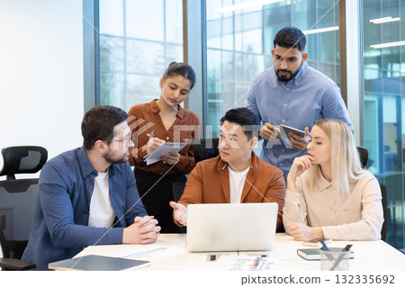 A business woman in a yellow blouse and hijab sits at her desk in an office, holding documents and a pen. 132335692