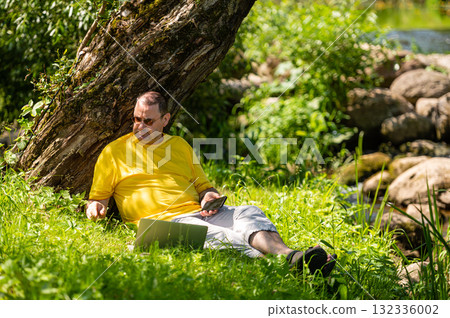 man with laptop and smartphone working outside in the meadow by the river, outdoor office concept 132336002