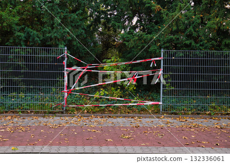Metal Fence Wrapped in Red and White Caution Tape Blocking Path Amidst Green Trees and Fallen Autumn Leaves on Paved Ground 132336061