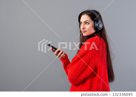 positive girl brunette in red sweater listens to music on smartphone in headphones, isolated on gray positive girl brunette in red sweater listens to music on smartphone in headphones, isolated on gray 132336095