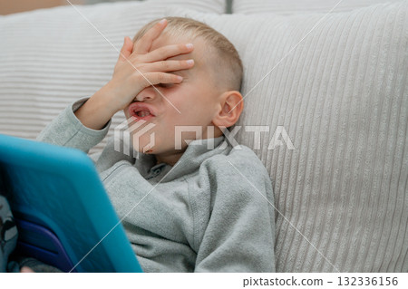 Young boy sitting on a couch with a tablet, covering his face with his hand in a playful facepalm candid moment of frustration Young boy sitting on a couch with a tablet, covering his face with his hand in a playful facepalm candid moment of frustration 132336156