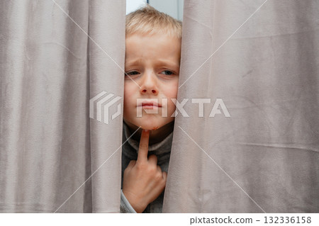 Boy with finger on chin looks thoughtful while standing between curtains calm indoor portrait Boy with finger on chin looks thoughtful while standing between curtains calm indoor portrait 132336158