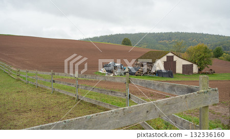 A rural barn surrounded by freshly plowed farmland and hills under a cloudy autumn sky. Peaceful countryside view with soft natural light and rustic atmosphere 132336160