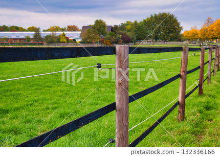 Serene Autumn Pasture Fence in Lush Green Field with Wooden Posts and Electric Wires Overlooking Distant Farm Buildings and Colorful Fall Trees 132336166