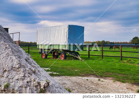 Vintage Green Farm Trailer Parked Beside White Lime Pile in Grassy Field with Wooden Fence and Distant Grazing Horses Under Blue Autumn Sky 132336227