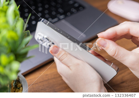Hands precisely using a SIM ejector tool to access the card slot of a sleek, modern smartphone, demonstrating essential mobile network configuration 132336407