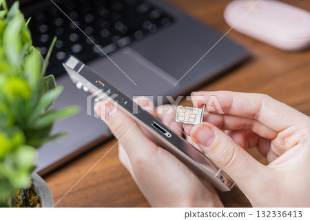 Human hands inserting a micro SIM card into a modern silver smartphone, symbolizing mobile network connectivity and essential telecommunication device setup on a busy desk 132336413