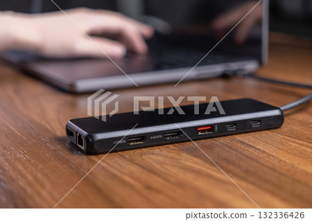 Black USB-C multiport hub connected to a laptop on a wooden desk, offering expanded digital connectivity and boosting productivity in modern work 132336426