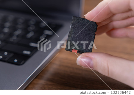 Person's hand holding a black digital SD memory card for versatile data storage and transfer, with a blurred laptop in a modern technology context Person's hand holding a black digital SD memory card for versatile data storage and transfer, with a blurred laptop in a modern technology context 132336442