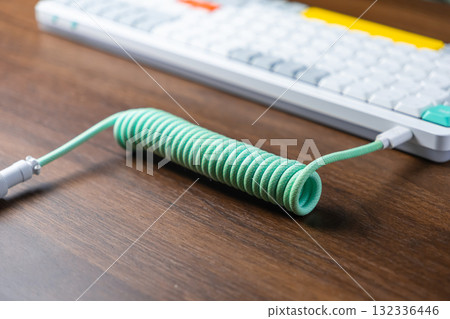 Mint green coiled braided USB cable connected to a modern mechanical keyboard on a wooden desk, enhancing desktop aesthetics for work or gaming Mint green coiled braided USB cable connected to a modern mechanical keyboard on a wooden desk, enhancing desktop aesthetics for work or gaming 132336446