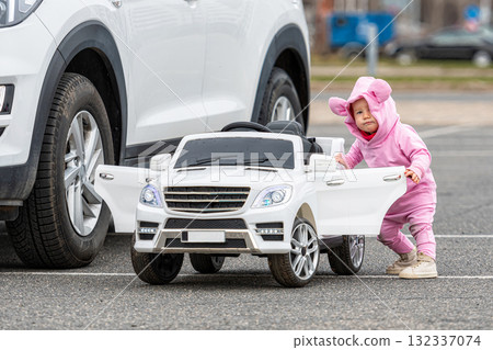 a little girl at a big white childrens electric toy car in the parking lot next to a real car 132337074