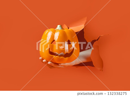 Hand Holding Jack-O'-Lantern. Hand reaches through torn orange paper holding a plastic Jack-O'-Lantern candy bucket. Hand Holding Jack-O'-Lantern. Hand reaches through torn orange paper holding a plastic Jack-O'-Lantern candy bucket. 132338275