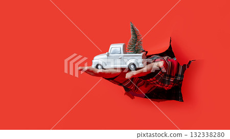Merry Christmas. Hands in plaid sleeves hold white toy truck carrying mini Christmas tree through torn red background. Merry Christmas. Hands in plaid sleeves hold white toy truck carrying mini Christmas tree through torn red background. 132338280
