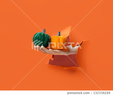 Hand holding soft wool pumpkin with golden stem through torn orange paper background. Hand holding soft wool pumpkin with golden stem through torn orange paper background. 132338284