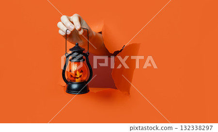 A hand holds a Halloween lantern with glowing Jack-O'-Lantern face through a ripped orange paper wall. 132338297