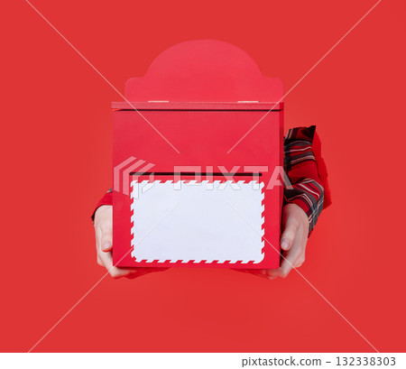 Merry Christmas, hand holds red holiday mailbox with an envelope inserted, festive plaid shirt visible through torn paper background. 132338303
