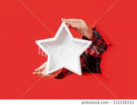 Hands in plaid shirt hold white star-shaped tray emerging through torn red paper, against festive red background. 132338310