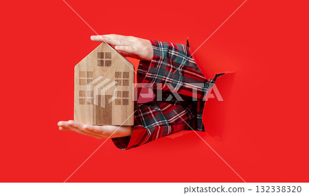 Merry Christmas. hand holding wooden toy house and pointing at it through torn red paper, plaid shirt visible. Merry Christmas. hand holding wooden toy house and pointing at it through torn red paper, plaid shirt visible. 132338320