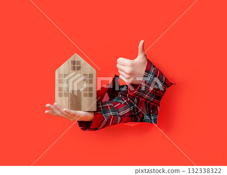 Merry Christmas, hands in plaid shirt hold wooden house while giving thumbs-up gesture through torn red paper. 132338322