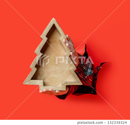 Merry Christmas, hand in plaid shirt holds a wooden tray shaped like a Christmas tree through a torn red paper background. Merry Christmas, hand in plaid shirt holds a wooden tray shaped like a Christmas tree through a torn red paper background. 132338324