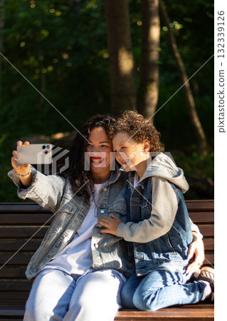 Mother making selfie together with her son. Taking selfie on smartphone. Smiling happy young family make photos outside in park to memorize moment. 132339126