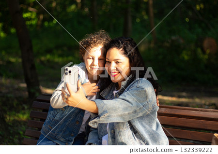 Young mother making selfie or video call with her son sitting on bench in park. Mom and boy calling grandparents. Concept of: connection, distance, technology 132339222