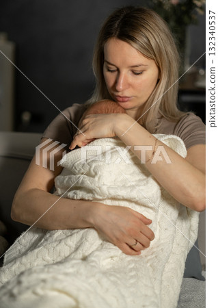 Mom lovingly holds her wrapped baby in a cozy living room, showcasing a tender bonding moment during a quiet afternoon Mom lovingly holds her wrapped baby in a cozy living room, showcasing a tender bonding moment during a quiet afternoon 132340537