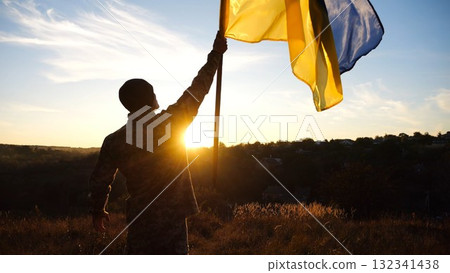 Young soldier of ukrainian army lifted blue-yellow banner in honor of the victory against russian aggression at sunset. Male military in uniform raised a waving flag of Ukraine at countryside Young soldier of ukrainian army lifted blue-yellow banner in honor of the victory against russian aggression at sunset. Male military in uniform raised a waving flag of Ukraine at countryside 132341438