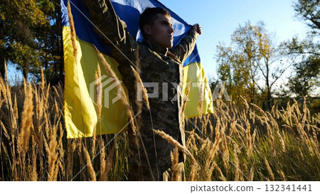 Military man in uniform stands with raised over head flag of Ukraine at countryside. Male ukrainian army soldier with lifted national banner in honor of victory against russian aggression. End of war 132341441
