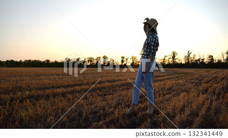 Adult agronomist in straw hat exploring barley on meadow at sunset. Female farmer examining ripe wheat at field on dusk. Beautiful scenic landscape. Concept of agriculture and agronomy business Adult agronomist in straw hat exploring barley on meadow at sunset. Female farmer examining ripe wheat at field on dusk. Beautiful scenic landscape. Concept of agriculture and agronomy business 132341449