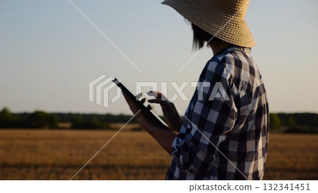 Female agronomist in straw hat monitoring harvest on barley field at sunset. Adult farmer using digital tablet on wheat meadow at dusk. Beautiful scenic landscape. Concept of agricultural business 132341451