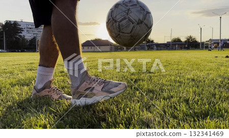 Male feet of professional footballer kicking ball at green field. Legs of young man juggling soccer ball on stadium at sunset. Sportsman practicing tricks outdoor. Freestyle football concept 132341469