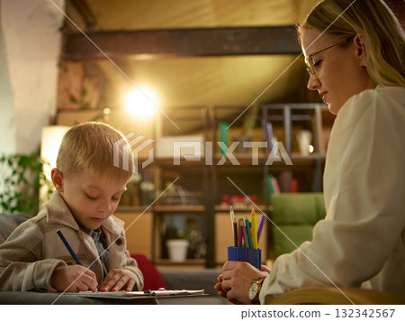 Child drawing with pencil during therapy session while psychologist observes attentively Child drawing with pencil during therapy session while psychologist observes attentively 132342567