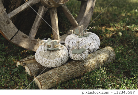 Handmade lace fabric pumpkins rest on cut logs near old wooden wagon wheel, rustic outdoor garden scene with cozy autumn mood 132343597