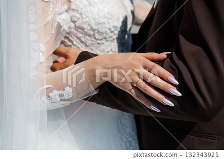 Wedding Day Close-Up: Bride and Groom Hands with Rings, White Lace Dress, Dark Suit, Romantic Embrace, Sunlight, Celebration, Intimate Moment. 132343921