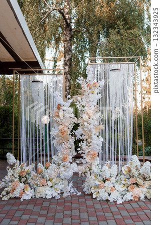 Romantic Wedding Ceremony Backdrop with White Floral Arrangement, String Curtains, Golden Accents, and Outdoor Garden Setting at Dusk. 132343925