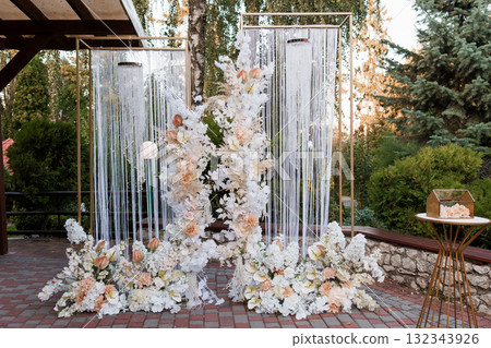 Romantic Outdoor Wedding Ceremony Backdrop with Floral Arch, White Curtains, and Golden Accents under Lush Green Trees at Dusk. 132343926