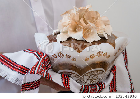 Traditional Ukrainian Easter Bread Paska with Floral Decorations Held in Hands, White Shirt, Red Embroidery, Festive Spring Holiday Still Life. Traditional Ukrainian Easter Bread Paska with Floral Decorations Held in Hands, White Shirt, Red Embroidery, Festive Spring Holiday Still Life. 132343936