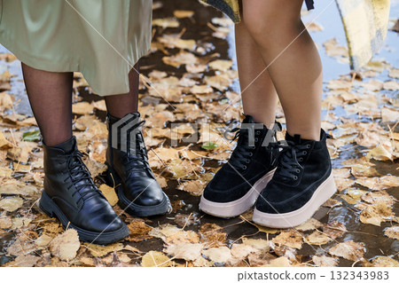 Autumn Fashion Boots Two Womens Legs and Black Leather Ankle Boots Standing in Fallen Leaves, Outdoor Style, Fall Colors, Close-Up Footwear. 132343983
