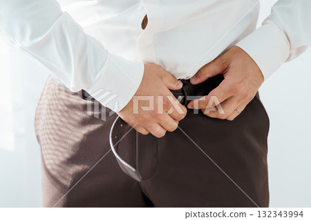 Man Fastening Brown Leather Belt with Silver Buckle. Business Attire. Waist Up. Neutral Tones. Formal Style. Studio Shot. Professionalism. Man Fastening Brown Leather Belt with Silver Buckle. Business Attire. Waist Up. Neutral Tones. Formal Style. Studio Shot. Professionalism. 132343994