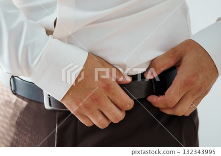 Man Fastening Brown Leather Belt with Silver Buckle Over Gray Trousers. Formal Attire, Business Style, Close-Up Shot, Dark Tone. 132343995