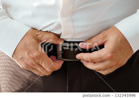 Groom Fastening Brown Leather Belt with Black Buckle, Wedding Preparation, Close-Up of Man's Hands, Formal Attire, Warm Tones. 132343996