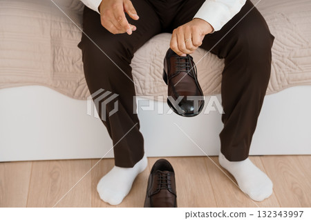 Man Tying Brown Leather Dress Shoes on Bed, White Socks, Formal Wear, Indoor Scene, Natural Light, Stylish Lifestyle, Neutral Bedroom Decor. Man Tying Brown Leather Dress Shoes on Bed, White Socks, Formal Wear, Indoor Scene, Natural Light, Stylish Lifestyle, Neutral Bedroom Decor. 132343997