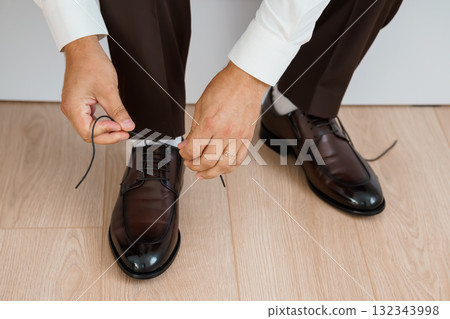Man Tying Shoelaces: Brown Leather Shoes, Formal Attire, Wooden Floor, Close-Up, Business Style, Polished Elegance, Preparation, Detail Shot. 132343998