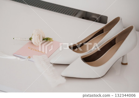 Elegant White Wedding Shoes with Delicate Rose and Lace Detail on a Light Surface, Romantic Bridal Preparation Scene. 132344006