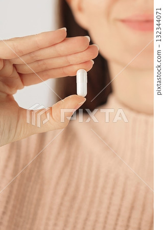 Vertical close-up of a hand holding a white capsule medication or vitamin. Concept of treatment, prevention and daily healthcare routine. Vertical close-up of a hand holding a white capsule medication or vitamin. Concept of treatment, prevention and daily healthcare routine. 132344071
