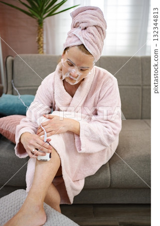 Woman shaving legs and wearing a face mask at home Woman shaving legs and wearing a face mask at home 132344813