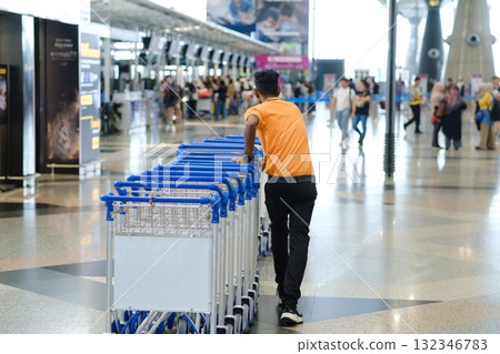 Man pushes luggage carts through busy airport terminal during daytime as travelers move around and check in for their flights Man pushes luggage carts through busy airport terminal during daytime as travelers move around and check in for their flights 132346783