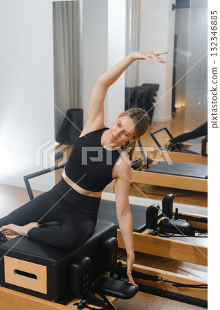 Fitness enthusiast practicing a side stretch on a reformer in a bright studio during a midday Pilates session Fitness enthusiast practicing a side stretch on a reformer in a bright studio during a midday Pilates session 132346885