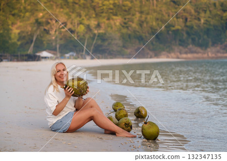 A woman enjoys a tropical beach holding a coconut in her hands, sitting at dawn next to fresh coconuts on a quiet coastline. Redang Island. Malaysia A woman enjoys a tropical beach holding a coconut in her hands, sitting at dawn next to fresh coconuts on a quiet coastline. Redang Island. Malaysia 132347135